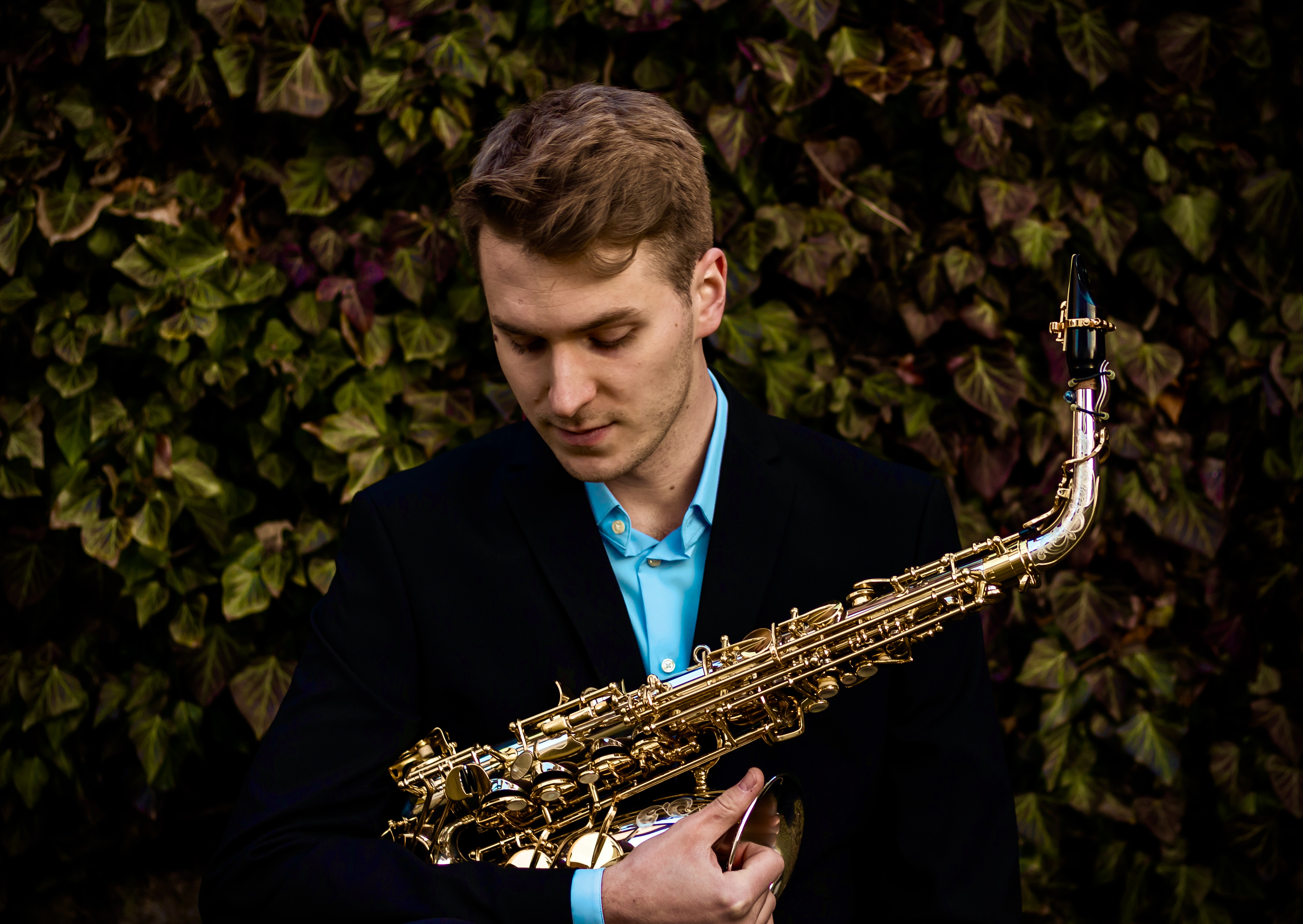 A young man with light brown hair, wearing a black suit jacket over a light blue collared shirt, holding a shiny golden sa...