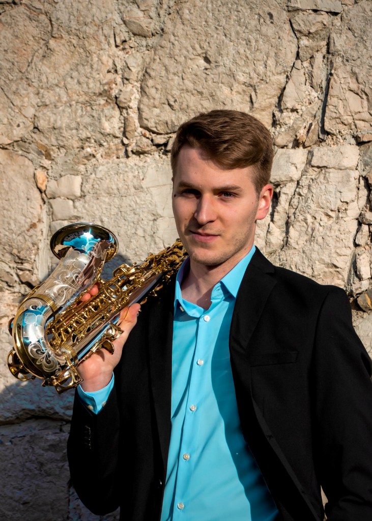 A man in a black suit, blue shirt and brown hair, holding a saxophone, in front of a stone wall.