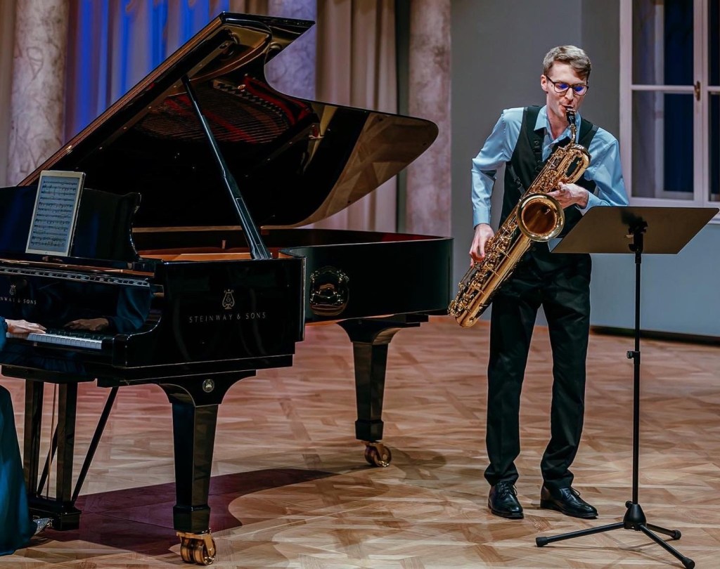 A male musician with glasses and a long-sleeved shirt plays a large gold saxophone at a grand piano during a concert, poss...