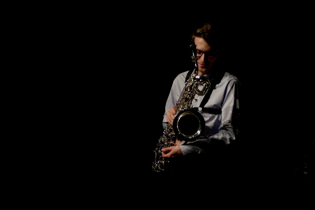 A man in a white shirt and dark glasses holds a saxophone in a dimly lit room.