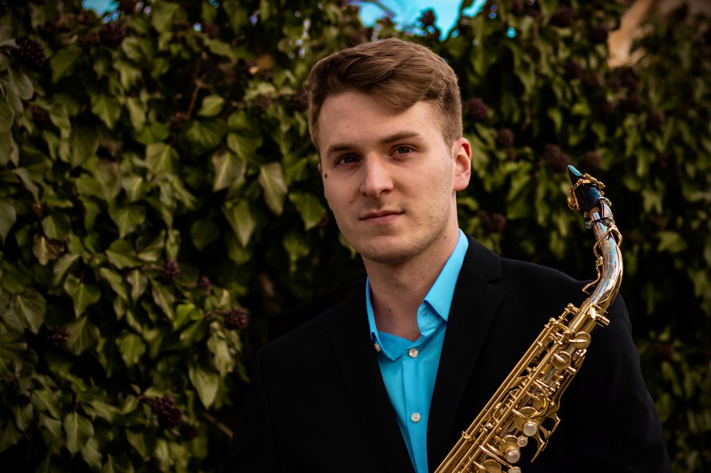 Young man in a black, business casual style blazer holding a saxophone, in front of dark green leaves on vines. A light-bl...