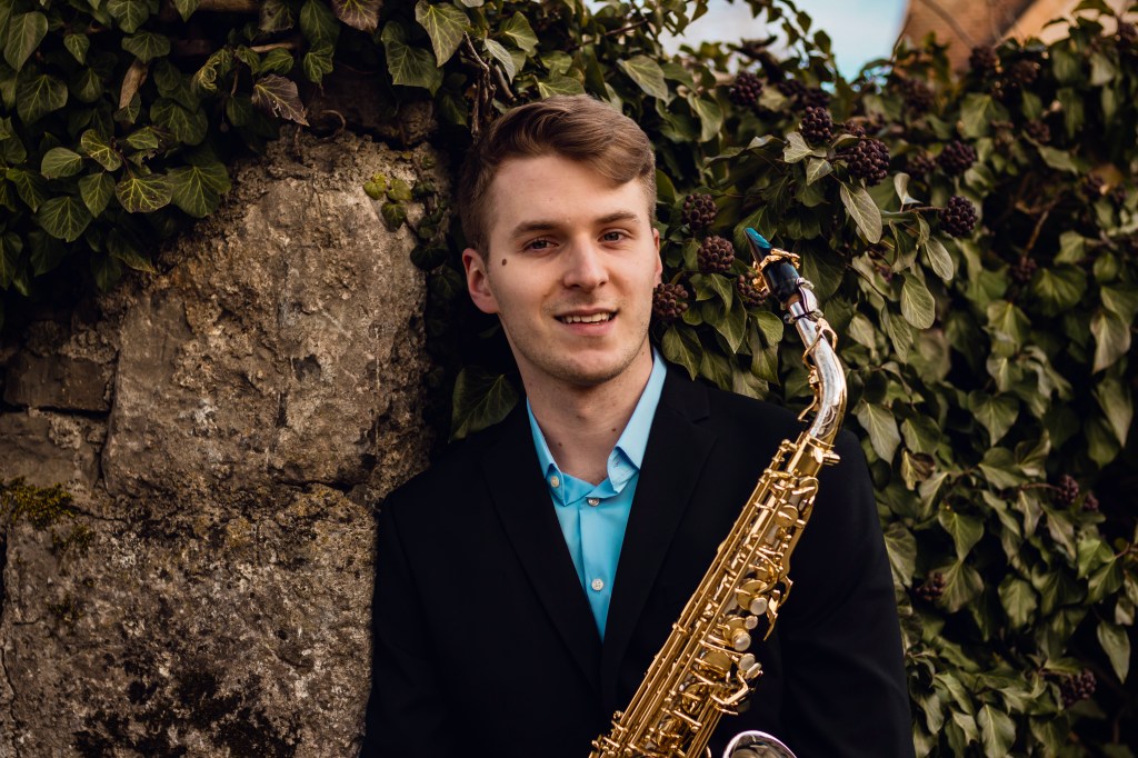 A young man in a suit, in front of a stone wall covered in English Ivy, and holding a golden saxophone, possibly for use a...