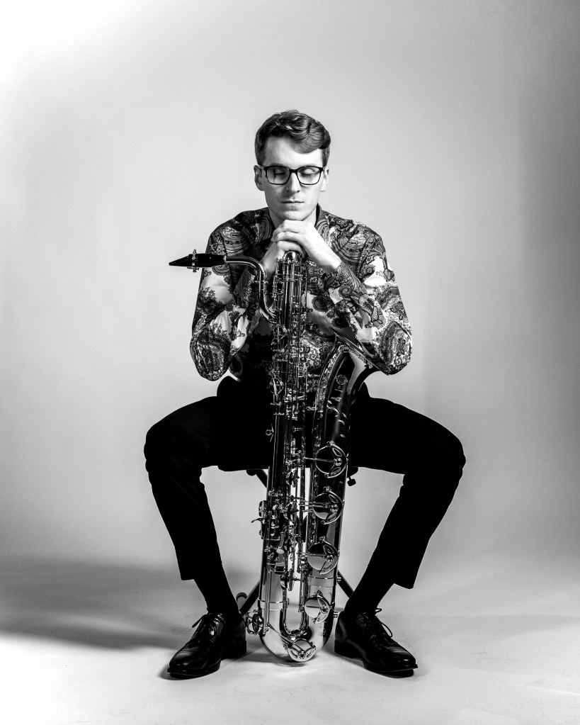 A man sitting on a chair with a saxophone leaning on this left knee, leaning against his right hand.