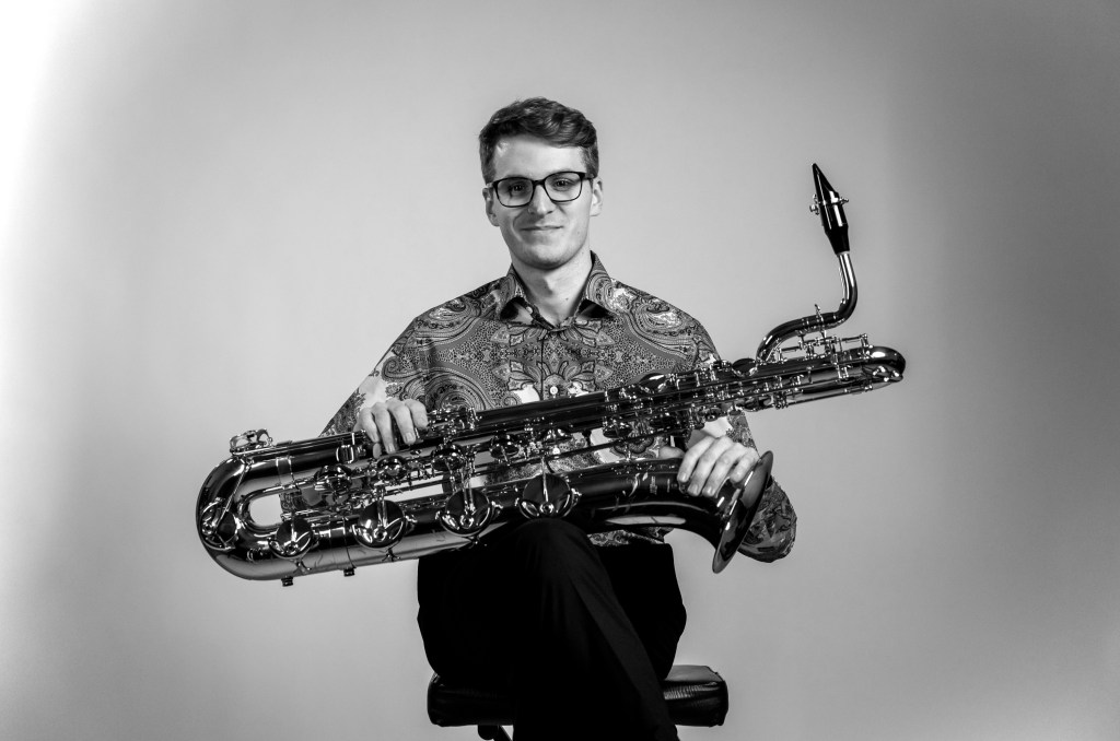 American man with short hair, a mustache, tie, and glasses holding a large trombone.