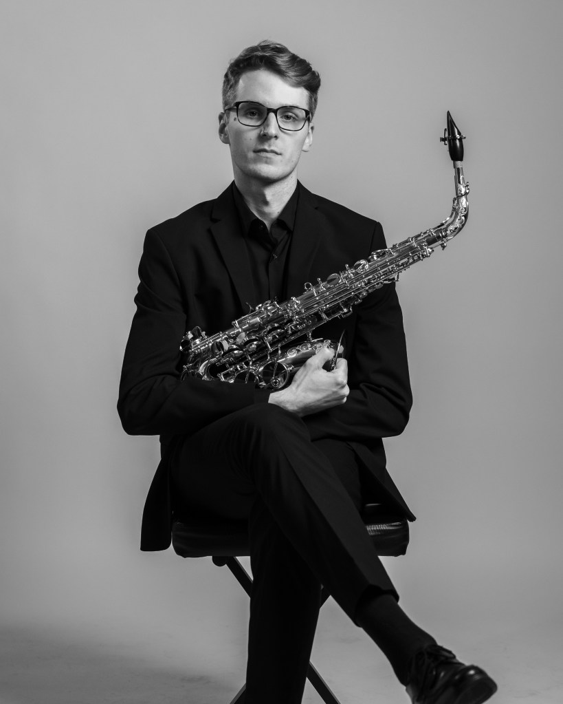 A black and white image of a man holding a saxophone, dressed in a dark suit and sitting on a stool.