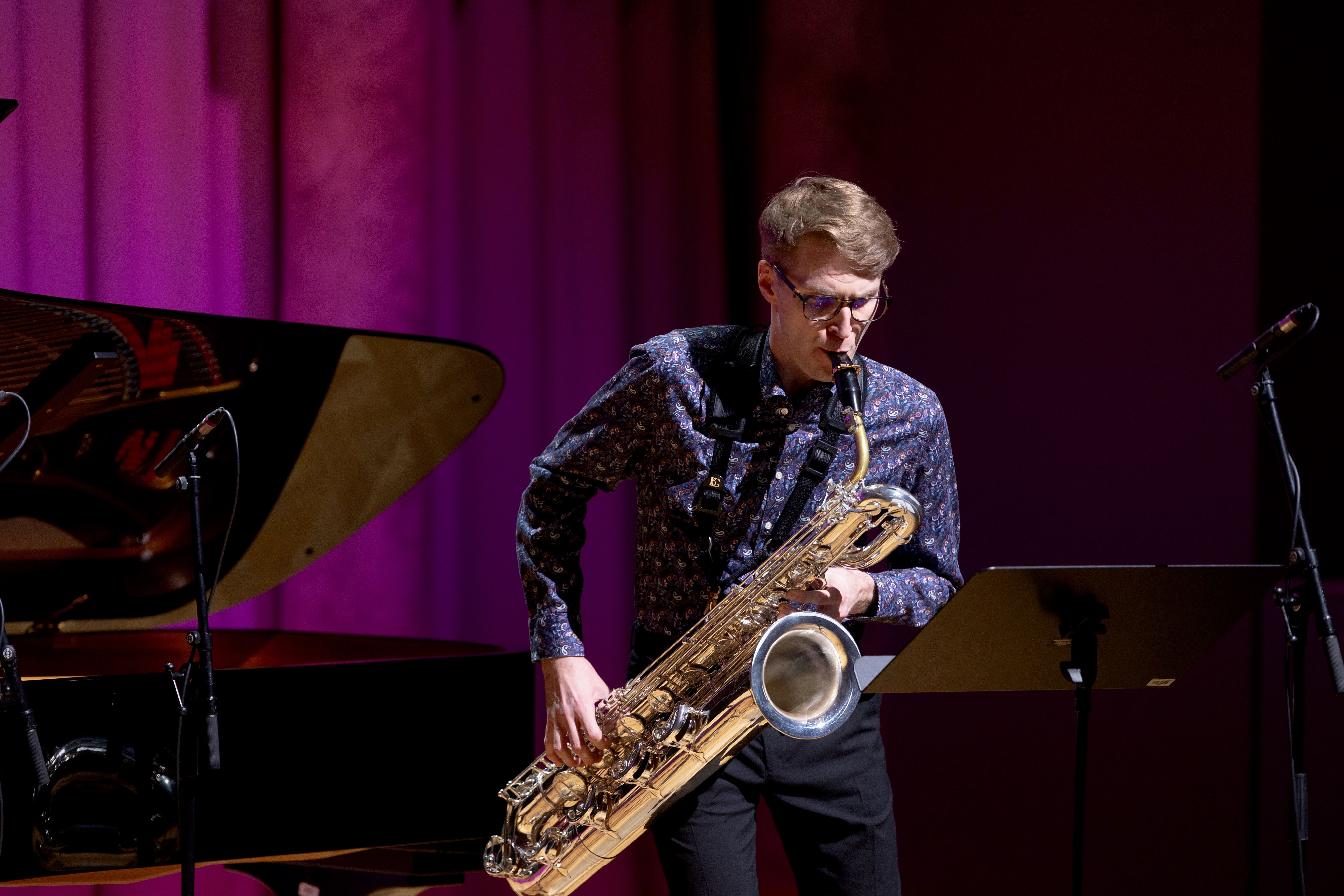 A man playing a low-frequency band instrument, possibly a baritone saxophone, with piano and microphone in the background,...